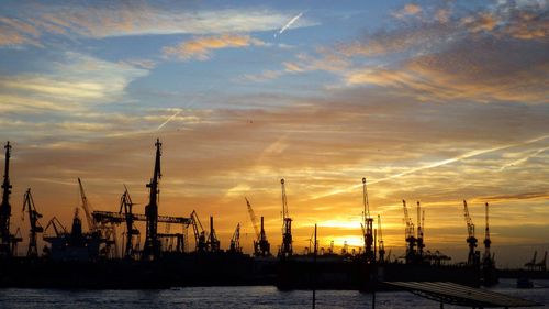 Silhouette cranes at commercial dock against sky at sunset