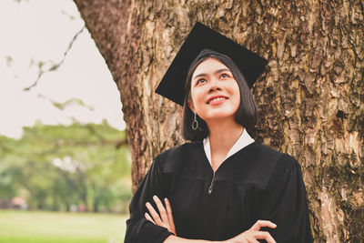 Thoughtful young woman in graduation gown with arms crossed standing by tree at park