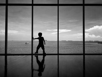 Silhouette boy looking at sea against sky through window