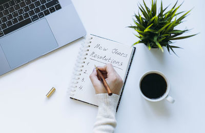 Notepad and woman hand writing new year resolutions. flower, laptop and coffee cup on background.