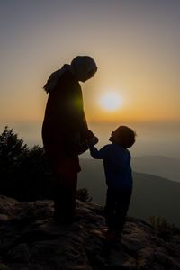Full length of mother and son holding hands while standing on mountain peak against sky during sunset