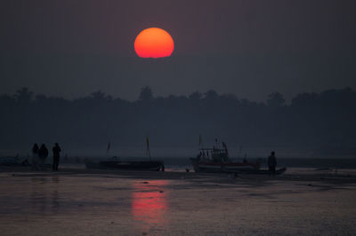 Silhouette people and boats at riverbank against sun in sky