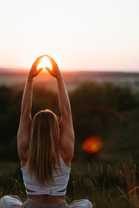 Back view on woman sitting in meditation yoga pose and catching sun by hands at sunset outdoors	
