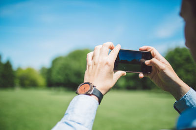 Cropped hands of man photographing green landscape through mobile phone during sunny day
