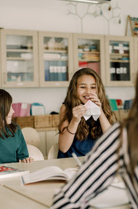 Smiling female student holding paper craft while sitting by table in classroom