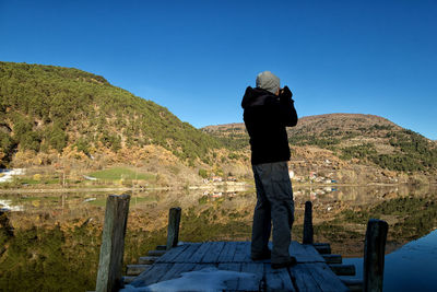 Man photographing lake by mountain while standing on pier against clear sky