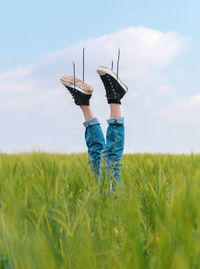 Upside down faceless person in denim and black sneakers raising legs out of green grass of field in summer