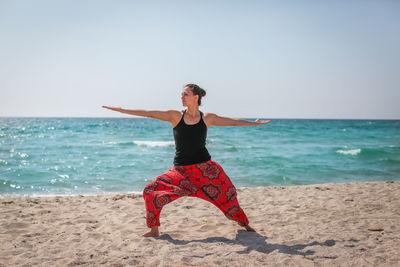 Mid adult woman exercising while standing at beach against clear sky during sunny day