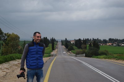 Smiling man holding camera on road against sky