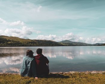 Rear view of men sitting by lake against sky