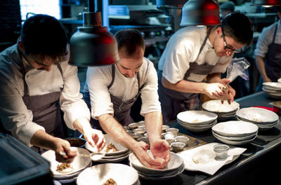 Young cooks men in aprons carefully serving meal in white ceramic dishes on restaurant kitchen