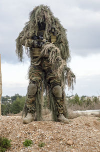 Army man in ghillie suit with rifle standing on land against sky