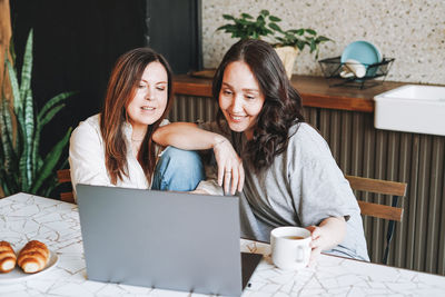 Adult smiling brunette middle aged women friends using laptop in kitchen at home