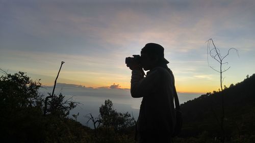 Side view of man photographing on mountain against sky during sunset