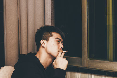 Young man smoking cigarette by window