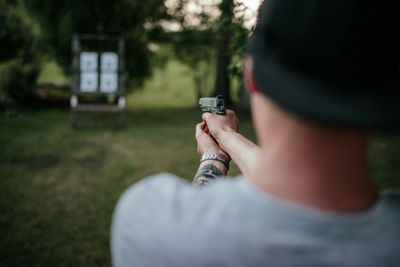 Young man with hat holding gun and shooting to target in countryside at daylight. rear view