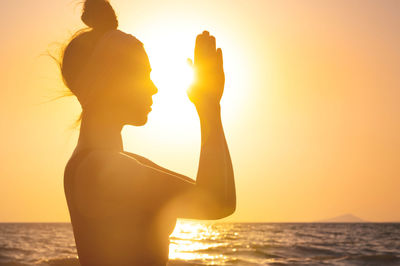 Rear view of woman with arms raised against sea during sunset