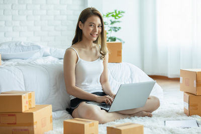 Full length of young woman using laptop while sitting by bed at home