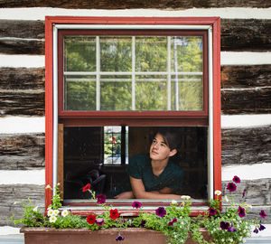 Teen boy looking out through window of rustic log cabin on summer day.