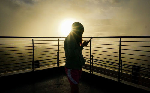 Woman standing by railing against sky during sunset