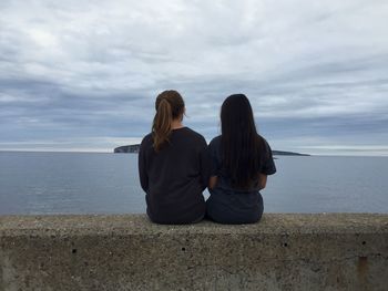 Rear view of women sitting on retaining wall against sea and cloudy sky