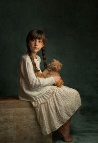 Girl dark hair two pigtails light dress sits legs tucked up holds favorite toy teddy bear in hands