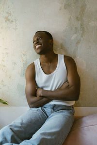 Smiling young man in undershirt sitting with arms crossed near wall at home