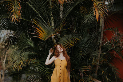 Carefree female with ginger hair and in summer dress standing under palm tree in exotic park in costa rica