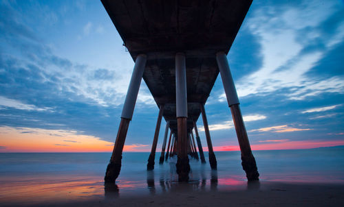 Below view of pier on shore at beach against sky during sunset