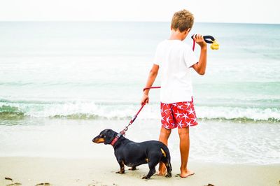 Rear view of boy with dog standing at beach against sky