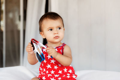 Cute baby girl holding sunglasses looking away while sitting on bed at home