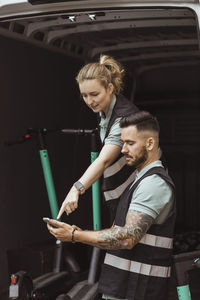 Young delivery woman pointing while male coworker using smart phone