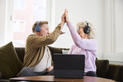 Senior man and woman giving each other high five while sitting in living room in front of digital tablet