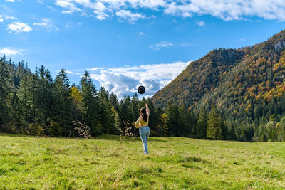 Full length of woman standing on field against sky, throwing hat in air.