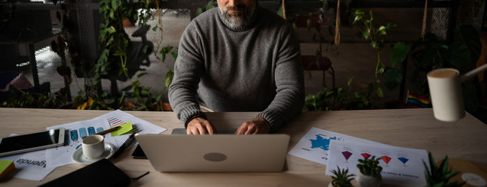 A man in a sitting position at a wooden table, using a laptop computer