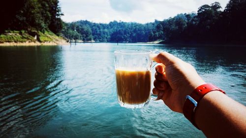 Close-up of hand holding drink by lake against trees