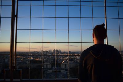 Rear view of young man looking through fence while standing against sky during sunset