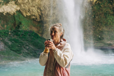 Portrait of an active woman drinking a hot drink in a mountain canyon near a large waterfall.