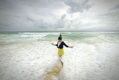 Playful woman enjoying on sea shore against cloudy sky