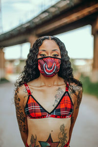 Portrait of young woman wearing mask standing against bridge