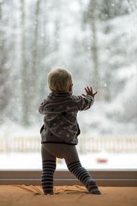 Rear view of baby boy looking through window while standing on bed at home