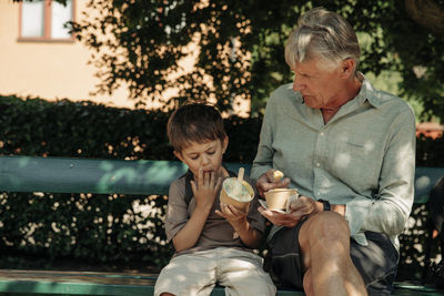 Grandfather and grandson eating ice cream in cups while sitting on bench