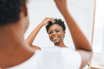 Portrait of beautiful young african american woman in white t-shirt makes hair style near mirror