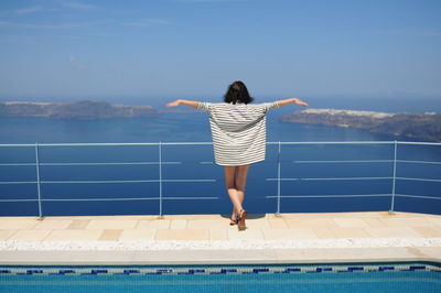 Rear view of woman standing with arms outstretched by railing at poolside against sea
