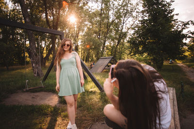 Young woman photographing her girlfriend in park during summer