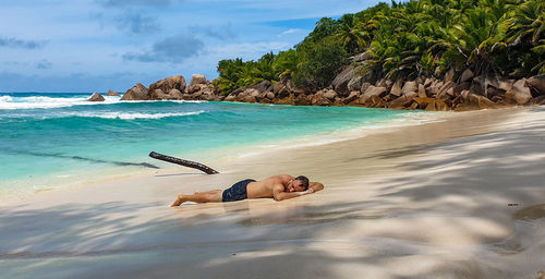 Young man in swim trunks laying on sandy beach on idyllic tropical island