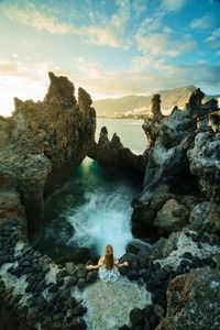 High angle view of young woman sitting on rock at beach against sky during sunset