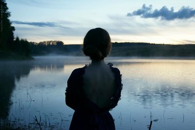 Rear view of woman standing at lakeshore against sky during sunset