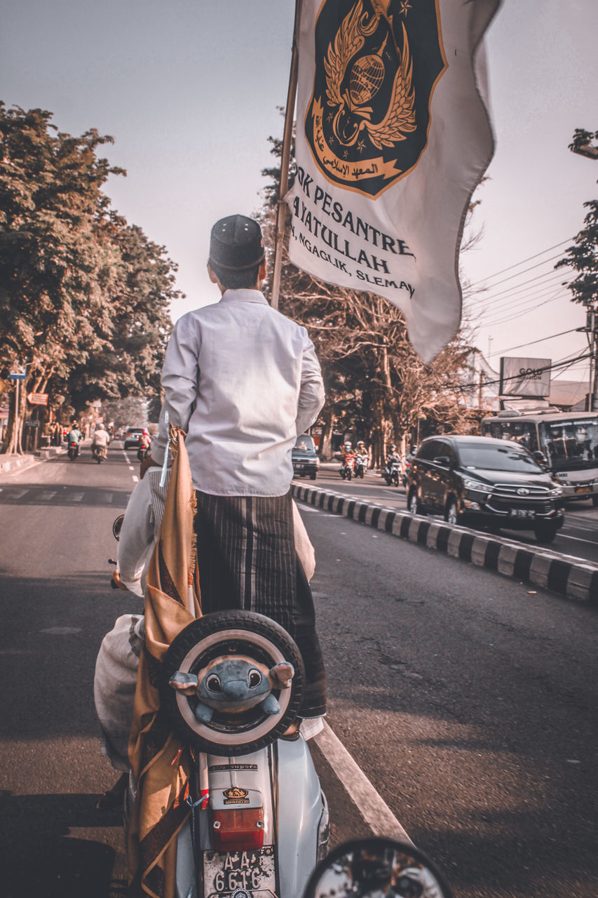 REAR VIEW OF MAN WALKING ON ROAD IN CITY