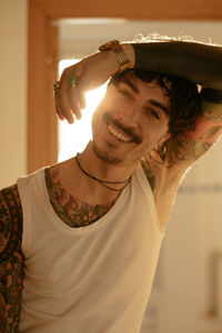 Young stylish guy with mustache and tattoos wearing white tank top and leaning against wall while looking at camera
